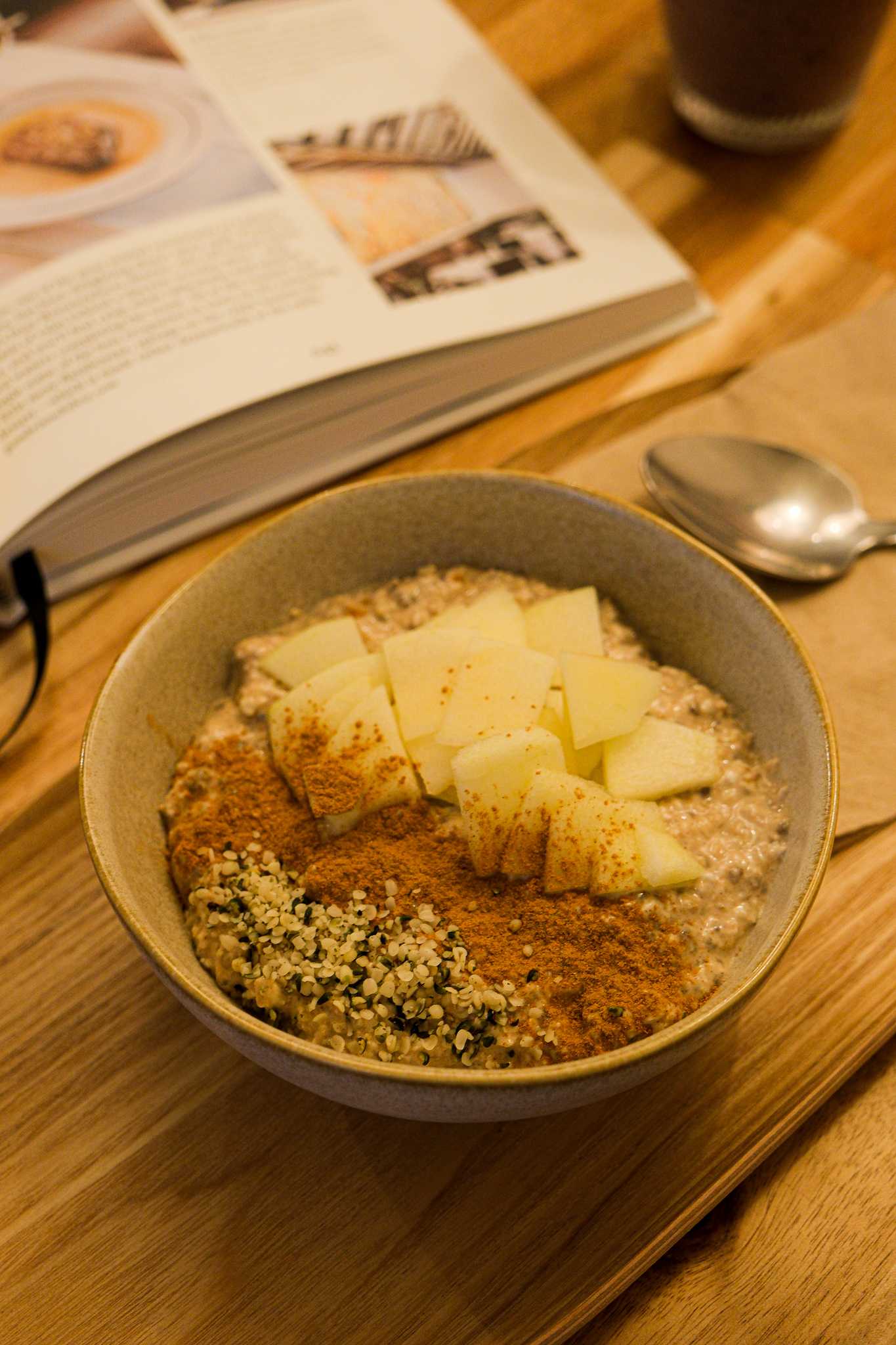 Uma visão aproximada de uma tigela de aveia coberta com fatias de maçã, sementes de cânhamo e canela, com um livro aberto e um copo de smoothie escuro ao fundo.
