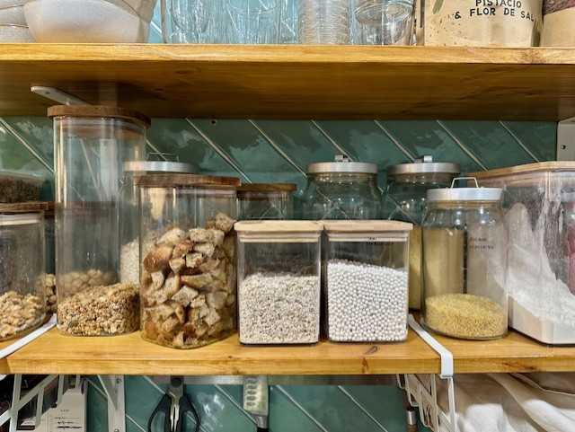 Tigela de papel vegware marrom contendo uma salada com ingredientes em cubos colocada em um banquinho redondo de madeira com pernas brancas contra uma parede de fundo creme.