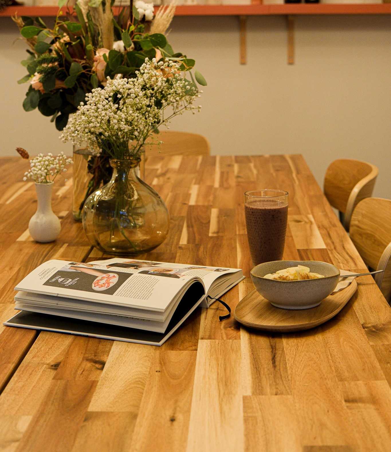 Uma mesa de café de madeira com um vaso de flores, um livro aberto, uma tigela de aveia coberta com frutas e um smoothie escuro em um copo.