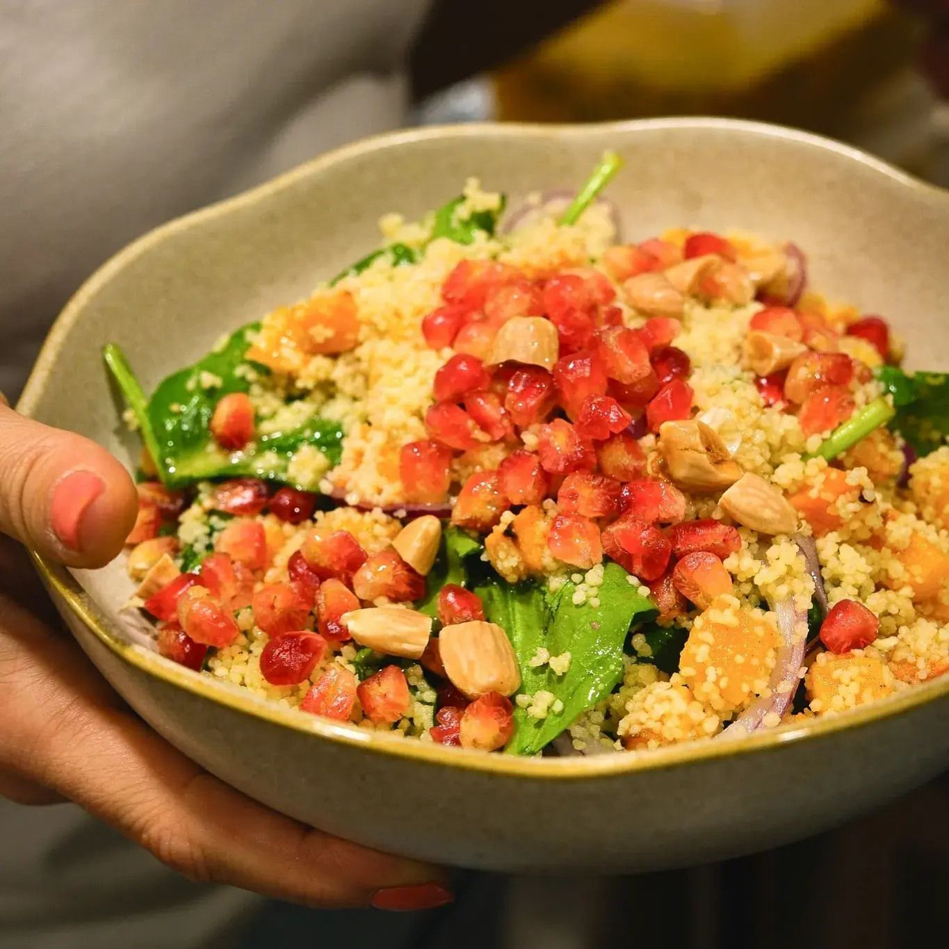 Um close-up de uma pessoa segurando uma tigela de salada colorida com espinafre, cuscuz, romã e amêndoas.