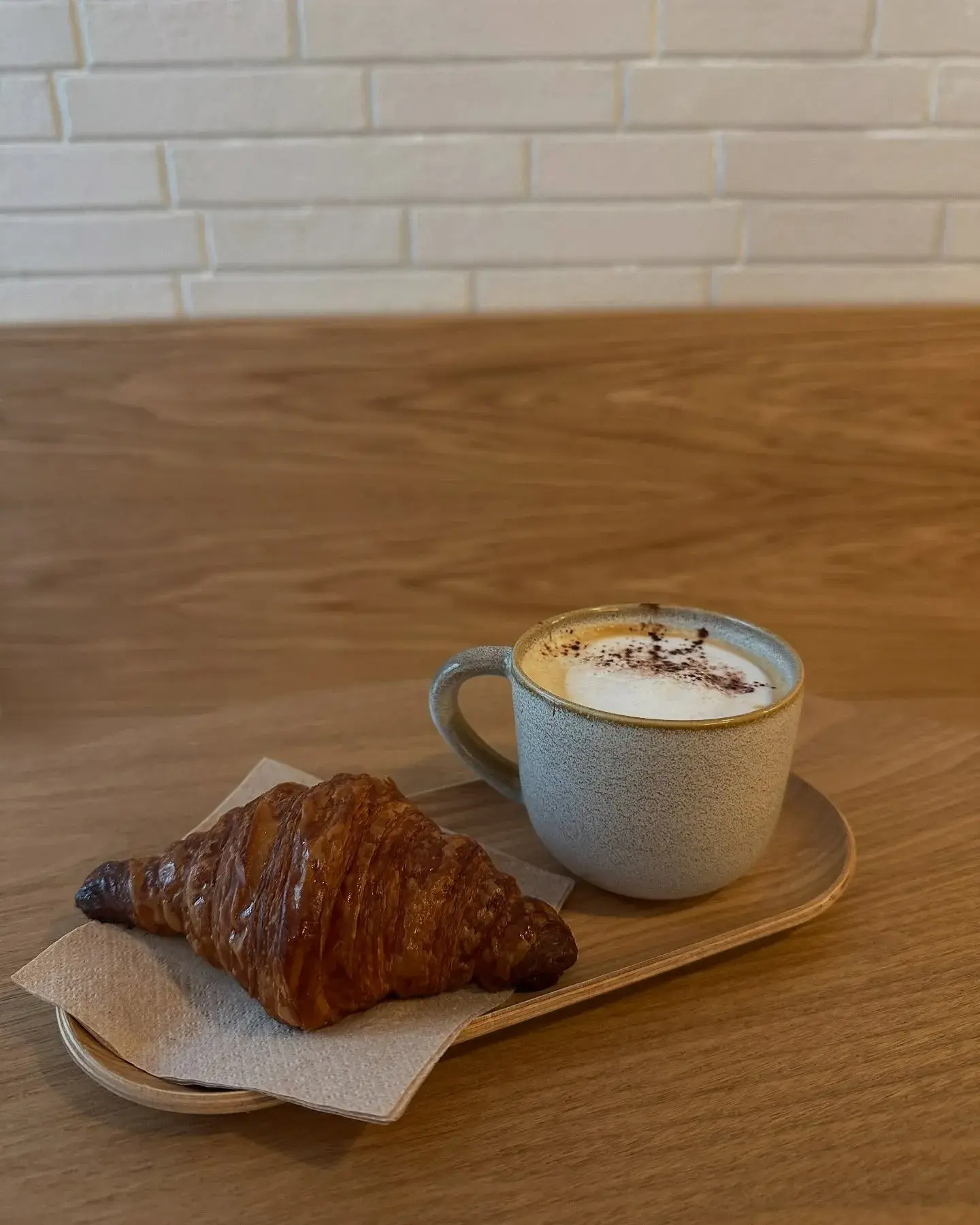 Um croissant amanteigado repousa sobre um guardanapo ao lado de um cappuccino espumoso em uma caneca texturizada.